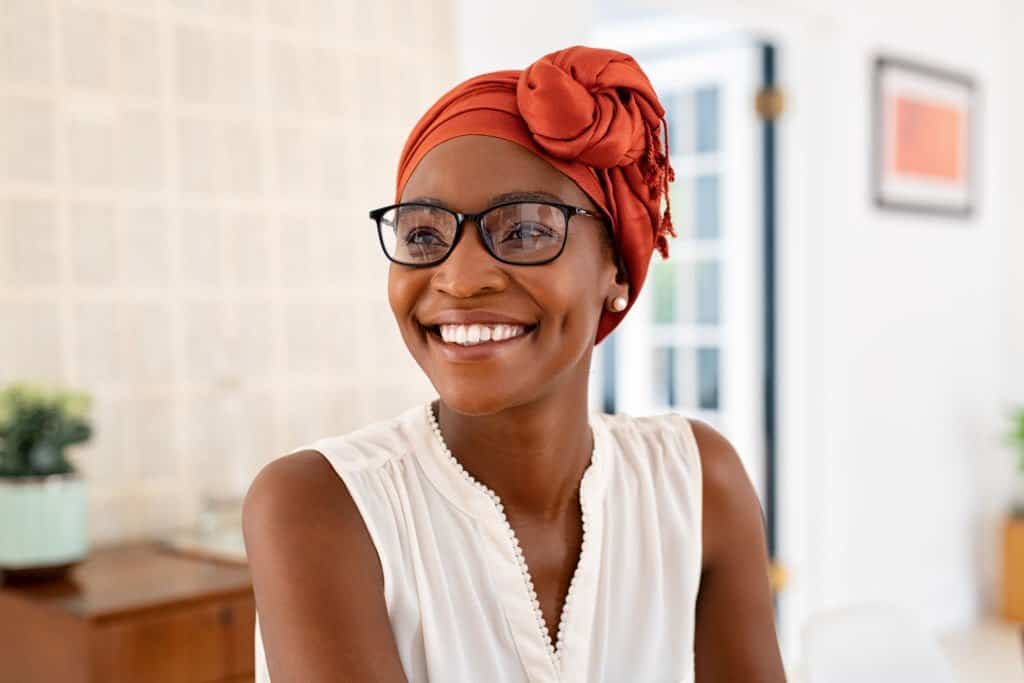 Cheerful mature black woman wearing spectacles and traditional turban at home. Beautiful african american woman with headscarf and eyeglasses sitting at home and looking away. Mature lady relaxing at home.